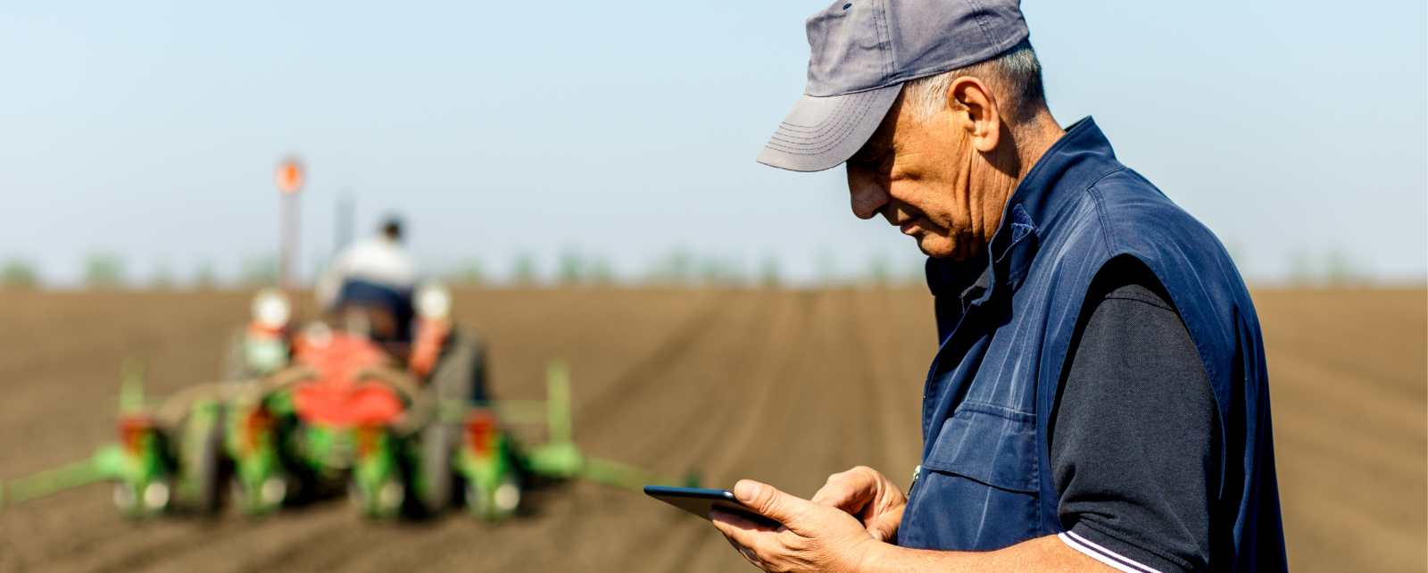 A farmer using a touchscreen tablet in a field, with another farmer operating heavy equipment in the